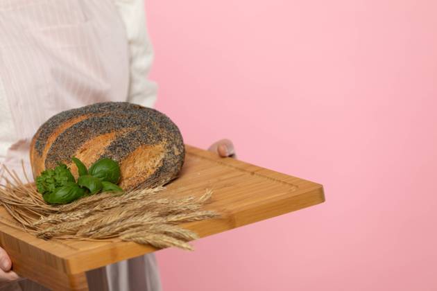 IMAGO Creative: Food, Mohn A young attractive saleswoman from a bakery shop holds a round loaf of bread sprinkled with poppy seeds and a pinch of fresh green herb leaves and rye ears on a wooden tray. The whole on a uniform light pink background. A young girl with a beautiful European beauty holds in her hands on a wooden board a freshly baked loaf of poppy seed bread, decorated with green herbs and ears of grain. The whole composition is set against a uniform light pink background. Copyright: xZoonar.com DANIELxDROBIKx 21500005