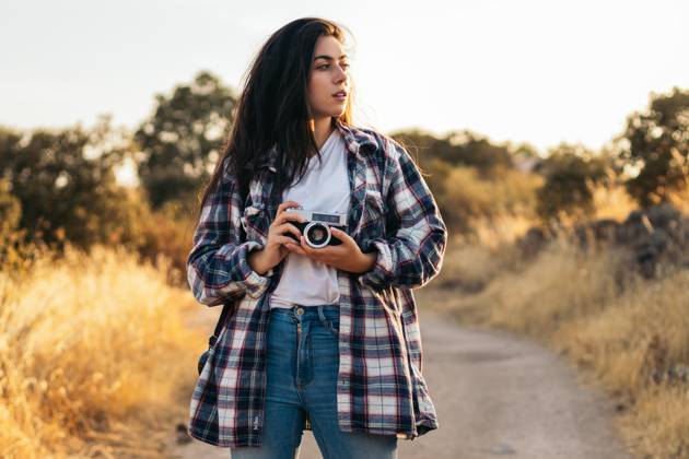 Young woman in checkered shirt and casual clothes taking photos with an analog camera in the field at sunset. The image conveys the concept of retro and vintage Model Released PedroMerino_ID19010_565591_011 Copyright: xx