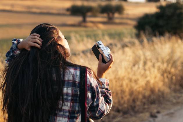 Young woman in checkered shirt and casual clothes taking photos with an analog camera in the field at sunset. The image conveys the concept of retro and vintage Model Released PedroMerino_ID19010_565592_012 Copyright: xx