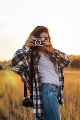 Young woman in checkered shirt and casual clothes taking photos with an analog camera in the field at sunset. The image conveys the concept of retro and vintage Model Released PedroMerino_ID19010_565598_016 Copyright: xx