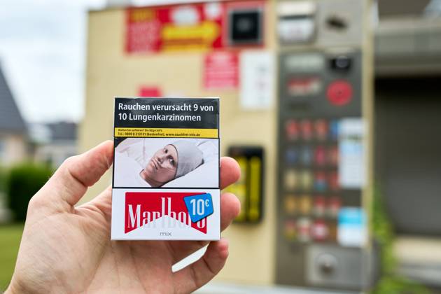 Hand hält eine Zigarettenschachtel von Marlboro zum Preis von 10 Euro vor einen Zigarettenautomat. Auf den Zigaretten ist ein Warnhinweis: Rauchen verursacht 9 von 10 Lungenkarzinomen