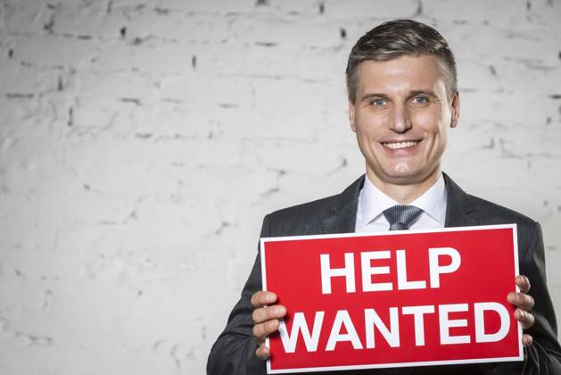 Portrait of smiling businessman holding help wanted sign while standing against white brick wall ,model released, Symbolfoto