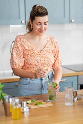culinary and people concept - happy smiling young woman making cocktail drinks with lime and peppermint at home kitchen. woman making cocktail drinks at home kitchen ,model released, Symbolfoto