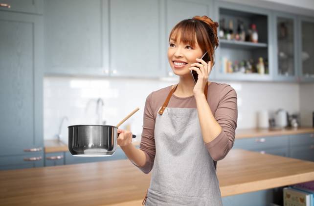culinary and people concept - happy smiling woman in apron with saucepan cooking food and calling on smartphone over kitchen background. happy female chef with saucepan calling on phone ,model released, Symbolfoto