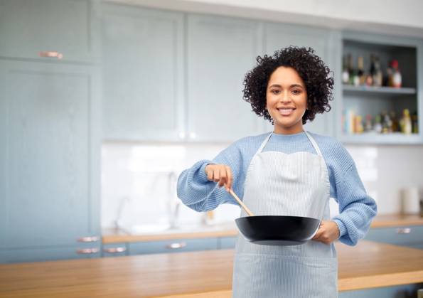 culinary and people concept - happy smiling woman in apron with frying pan and spoon cooking food over home kitchen background. happy woman cooking food in frying pan in kitchen ,model released, Symbolfoto
