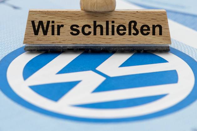 VW-Krise mit Gefahr der Werksschließung Ein Holzstempel ist beschriftet mit dem Aufdruck: Wir schließen, über einer VW-Aktie mit Logo - Symbolbild