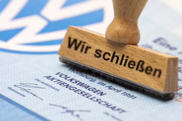 VW-Krise mit Gefahr der Werksschließung Ein Holzstempel ist beschriftet mit dem Aufdruck: Wir schließen, über einer VW-Aktie mit Logo - Symbolbild
