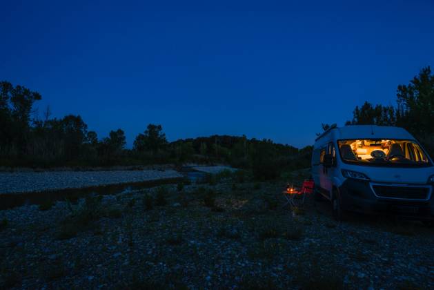 EIn Mann sitzt auf einem Campingstuhl vor einem Tisch an einem Sommerabend zur blauen Stunde vor seinem Camper. Deutschland Mit einem Campervan unterwegs