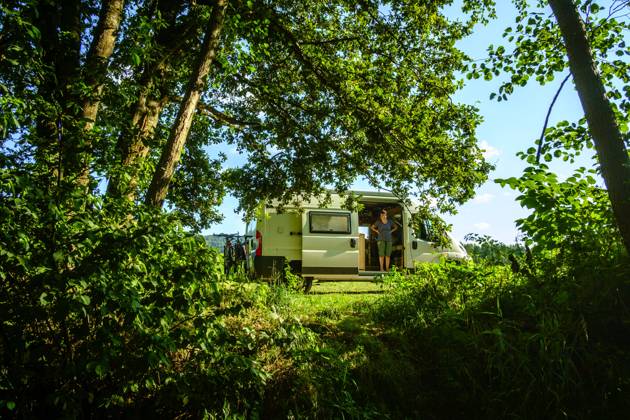 EIne Frau steht in der Schiebetür eines Campers, der in idyllischer Umgebung im Sommer auf einer Wiese zwischen Bäumen steht, Vogesen, Frankreich. Mit einem Campervan unterwegs