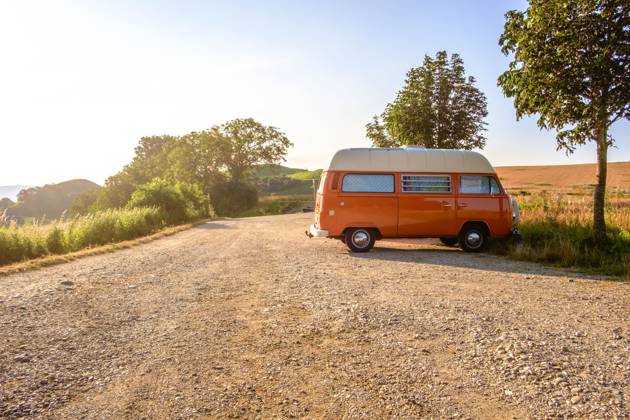 Ein Orange-weisser VW Bus Bulli steht auf einem Schotterparkplatz an einem Sommermorgen, Baden Württemberg, Deutschland. Ein Bulli steht in der Landschaft