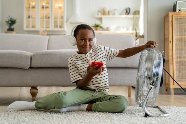 IMAGO Creative, Jahreszeit Sommer, Hitze Lächelnder, glücklicher afroamerikanischer Junge, der sich zu Hause vor einem elektrischen Ventilator entspannt Smiling happy African American boy child wearing headphones listening music or watching internet video while relaxing at home in front of electric fan. Children and summer heat ,model released, Symbolfoto Copyright: xZoonar.com DmitriixMarchenkox 22690747 ,model released, Symbolfoto ,property released