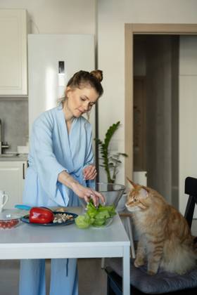 MR_ID27929_20617_TatyanaRangulova.jpg, PR_ID27929_10891_Cat.jpg, PR_ID27929_10892_Room.jpg A woman in a blue robe prepares vegetables at a kitchen table. A curious cat sits nearby, observing her actions. The scene captures a cozy, domestic moment., Property released Model Released LiliyaSolopova_ID27929_802060_006 Copyright: xLiliyaxSolopovax
