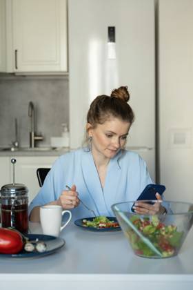 MR_ID27929_20617_TatyanaRangulova.jpg, PR_ID27929_10891_Cat.jpg, PR_ID27929_10892_Room.jpg A woman wearing a blue robe is sitting at a table in her kitchen, eating a fresh salad while looking at her smartphone. The scene conveys a relaxed domestic atmosphere., Property released Model Released LiliyaSolopova_ID27929_802062_007 Copyright: xLiliyaxSolopovax