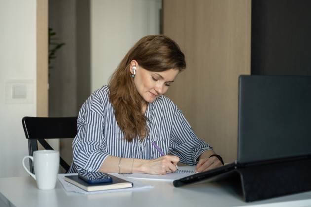 MR_ID27929_20617_TatyanaRangulova.jpg, PR_ID27929_10891_Cat.jpg, PR_ID27929_10892_Room.jpg A woman sits at a desk at home, writing in a notebook. She is wearing wireless earphones, a striped shirt, and is focused on her laptop. A mug and mobile phone are nearby., Property released Model Released LiliyaSolopova_ID27929_802068_011 Copyright: xLiliyaxSolopovax
