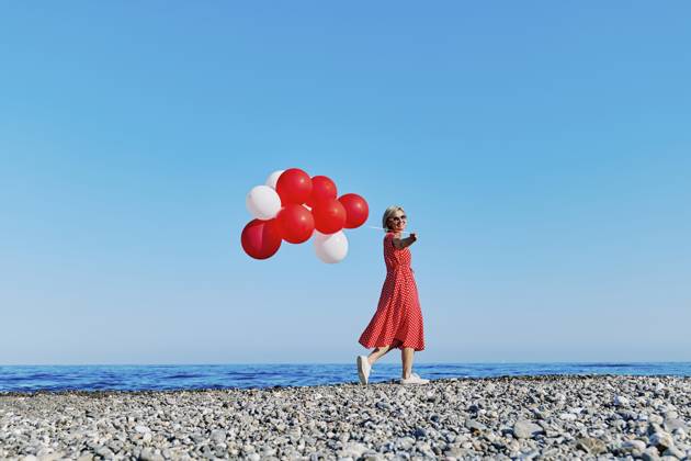 Blond woman in red polka dot dress holding balloons on a pebble beach by the Ionian Sea on a sunny day. model released, Symbolfoto, CATF00697