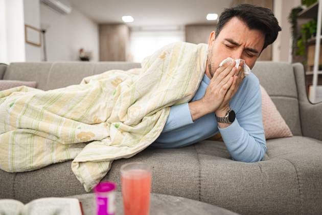 Sick man having a runny nose while lying on a sofa covered with blanket Man is seen laying on a couch, covering his face with a tissue, as he blows his nose due to sickness or allergies. His eyes may appear watery or tired., model released, , 63184974.jpg RECORD DATE NOT STATED