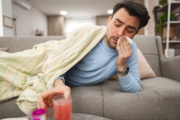Sick man having a runny nose while lying on a sofa covered with blanket Man is seen laying on a couch, covering his face with a tissue, as he blows his nose due to sickness or allergies. His eyes may appear watery or tired., model released, , 62041996.jpg RECORD DATE NOT STATED