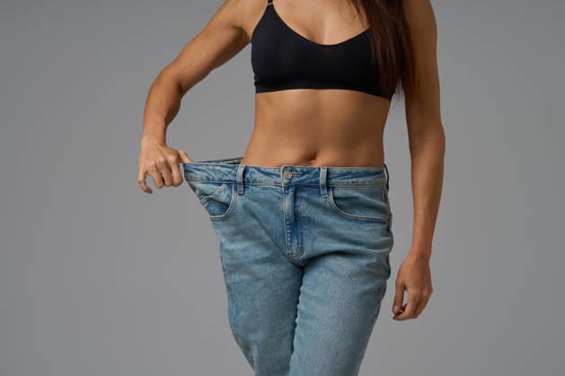 Fit Frau in schwarzem Sport-BH zieht lose Jeans, um Gewichtsverlust Fortschritte auf grauem Hintergrund zu zeigen Fit young woman wearing black sports bra and oversized jeans, pulling the waistband to display weight loss results. Studio shot on neutral gray backdrop. (License=RF) ,model released, Symbolfoto Copyright: xZoonar.com SergeyxMironovx 24323403 ,model released, Symbolfoto ,property released