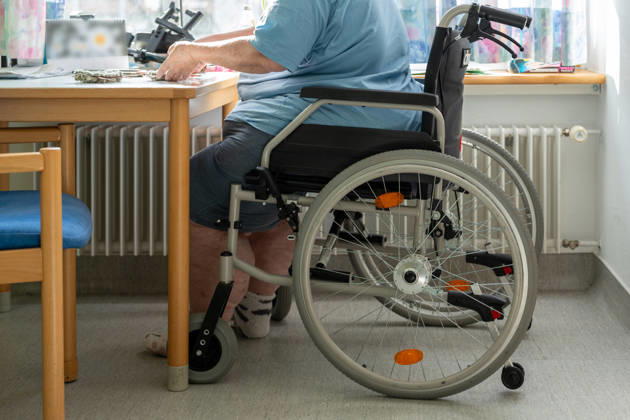 Ein älterer Mensch im Rollstuhl am Tisch verdeutlicht die Herausforderungen im Pflegealltag. Das Bild thematisiert Inklusion, Barrierefreiheit und die notwendige Unterstützung für ein würdevolles Leben.