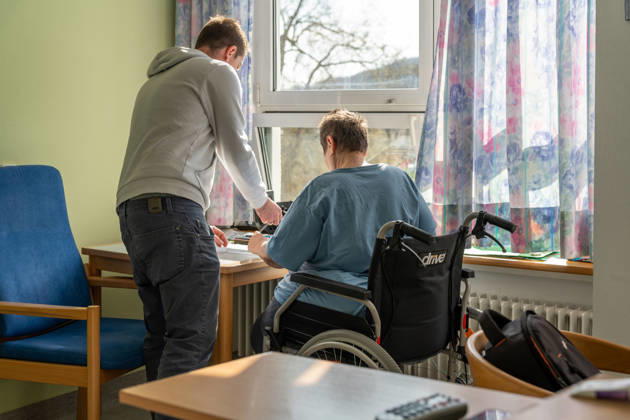Pfleger unterstützt einen Patienten im Rollstuhl bei einer Aufgabe am Tisch in einer Pflegeeinrichtung. Die Szene zeigt den Alltag in der Pflege, in dem Betreuung, Unterstützung und menschliche Fürsorge eine zentrale Rolle spielen.