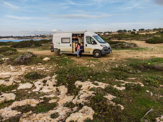 Camper sitzen in der Schiebetür ihres Campers auf einem unbefestigten Offroad Stellplatz an der Küste am Mittelmeer bei Tarent. Salento, Apulien, Italien. Ein Wohnmobil steht auf einem Schotterplatz am Ufer des Mittelmeers.
