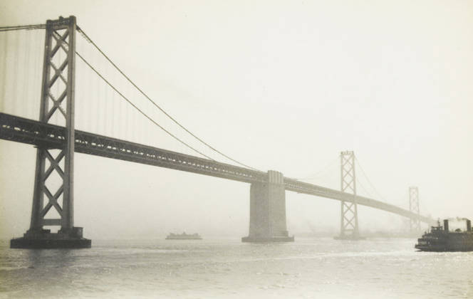 RECORD DATE NOT STATED Oakland Bridge, San Francisco, photograph, title: OAKLAND BRIDGE, SAN FRANCISCO , photographer: attributed to Wouter Cool, United States of America, 1936, North America, Attributed to, pasteboard, paper, gelatin silver print, developing gelatin silver print, height 150 mm x width 229 mm, height 315 mm x width 285 mm, bridge, A wide view of a suspension bridge spanning across a body of water, shown in a monochrome tone, with tall lattice bridge towers and diagonal suspension cables extending from the left foreground toward the right background, a central bridge pier rising from the water beneath the roadway, a small ship visible beneath the bridge near the center, and a ferry or boat at the right edge partially entering the frame, with the Copyright Penta Springs LCK260531_17994