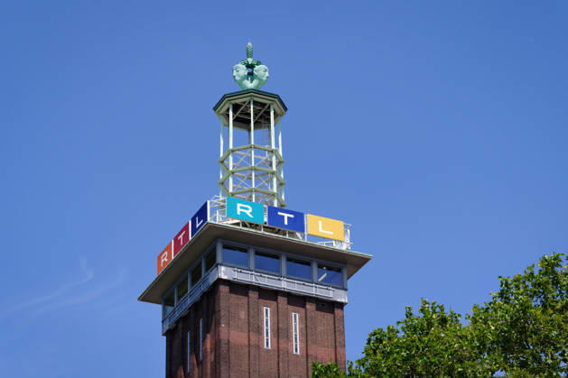 Detailaufnahme des Messeturms in Köln mit dem bunten RTL-Senderlogo und der markanten Turmspitze vor blauem Sommerhimmel mit grünen Blättern