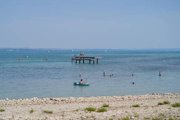 Bodensee: Badegäste und Wassersportler genießen einen sonnigen Sommertag im Bodensee. Menschen schwimmen im See, fahren mit Schlauchboot und Stand-up-Paddle; weitere Personen stehen auf einem Badesteg. Im Hintergrund Segelboote und das gegenüberliegende Ufer. Sommer, Hitze, Freizeit, Baden, Schwimmen, Wassersport, Tourismus, Ferien, Erholung, See, Strand, Badesteg, SUP, Panorama, Totale, Symbolbild. Copyright: xJOERANxSTEINSIEKx