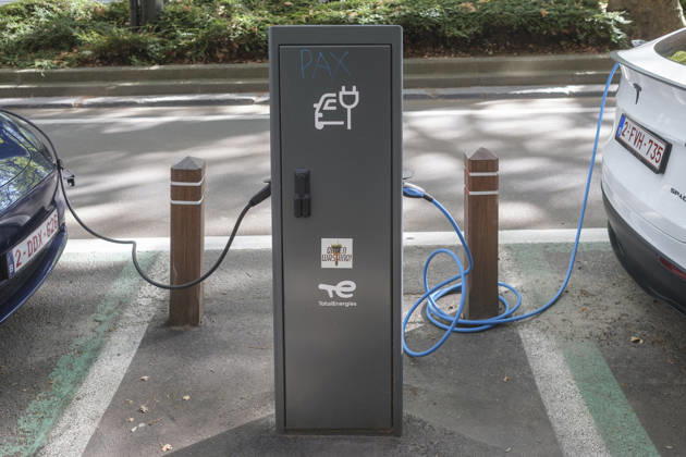 Zwei Elektrofahrzeuge sind über Ladekabel mit einer anthrazitfarbenen Ladesäule von TotalEnergies am Straßenrand verbunden. Auf der linken Seite ist das Heck eines blauen Fahrzeugs zu sehen, während rechts ein weißes Tesla-Modell mit belgischem Kennzeichen lädt. Die Ladesäule im Zentrum weist neben dem offiziellen Logo und einem E-Auto-Symbol ein handgemaltes Graffiti-Wort PAX sowie einen Aufkleber mit der Aufschrift Green Washing! auf.