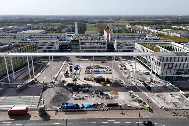 Umgestaltung des Willy-Brandt-Platzes an den Riem Arcaden: Versiegelte Flaechen werden in Gruenflaechen umgewandelt. Am Willy-Brandt-Platz vor den Riem Arcaden in Muenchen laufen die Bauarbeiten zur Neugestaltung. Im Zuge der Umgestaltung sollen bisher versiegelte Flaechen entsiegelt und durch Gruenflaechen ersetzt werden. Baustellenabsperrungen, Baumaterial und ein Radlader praegen derzeit das Bild des Platzes.