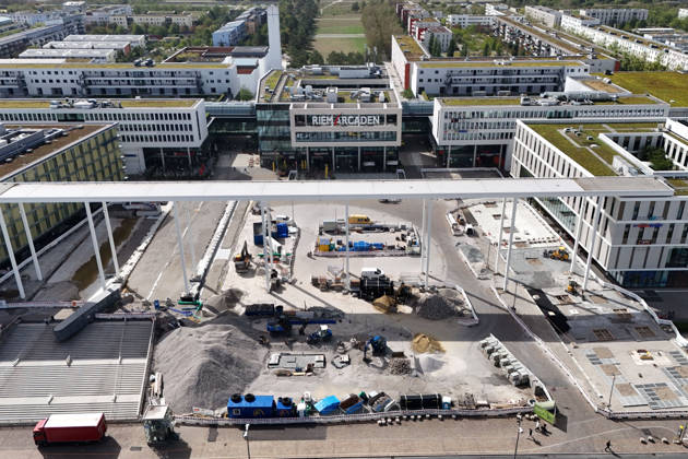 Umgestaltung des Willy-Brandt-Platzes an den Riem Arcaden: Versiegelte Flaechen werden in Gruenflaechen umgewandelt. Am Willy-Brandt-Platz vor den Riem Arcaden in Muenchen laufen die Bauarbeiten zur Neugestaltung. Im Zuge der Umgestaltung sollen bisher versiegelte Flaechen entsiegelt und durch Gruenflaechen ersetzt werden. Baustellenabsperrungen, Baumaterial und ein Radlader praegen derzeit das Bild des Platzes.