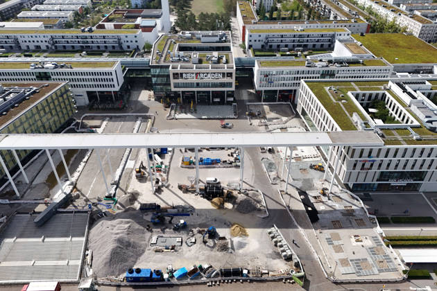 Umgestaltung des Willy-Brandt-Platzes an den Riem Arcaden: Versiegelte Flaechen werden in Gruenflaechen umgewandelt. Am Willy-Brandt-Platz vor den Riem Arcaden in Muenchen laufen die Bauarbeiten zur Neugestaltung. Im Zuge der Umgestaltung sollen bisher versiegelte Flaechen entsiegelt und durch Gruenflaechen ersetzt werden. Baustellenabsperrungen, Baumaterial und ein Radlader praegen derzeit das Bild des Platzes.