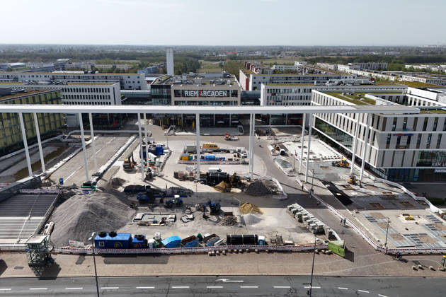 Umgestaltung des Willy-Brandt-Platzes an den Riem Arcaden: Versiegelte Flaechen werden in Gruenflaechen umgewandelt. Am Willy-Brandt-Platz vor den Riem Arcaden in Muenchen laufen die Bauarbeiten zur Neugestaltung. Im Zuge der Umgestaltung sollen bisher versiegelte Flaechen entsiegelt und durch Gruenflaechen ersetzt werden. Baustellenabsperrungen, Baumaterial und ein Radlader praegen derzeit das Bild des Platzes.
