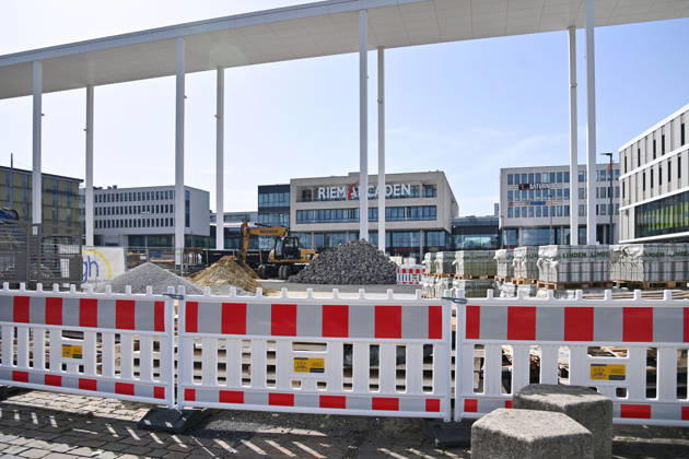 Umgestaltung des Willy-Brandt-Platzes an den Riem Arcaden: Versiegelte Flaechen werden in Gruenflaechen umgewandelt. Am Willy-Brandt-Platz vor den Riem Arcaden in Muenchen laufen die Bauarbeiten zur Neugestaltung. Im Zuge der Umgestaltung sollen bisher versiegelte Flaechen entsiegelt und durch Gruenflaechen ersetzt werden. Baustellenabsperrungen, Baumaterial und ein Radlader praegen derzeit das Bild des Platzes.
