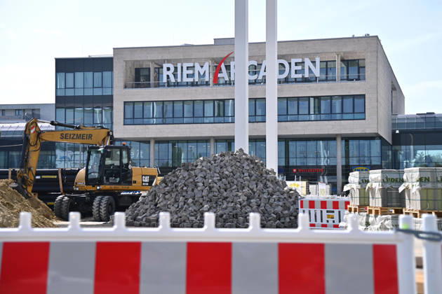 Umgestaltung des Willy-Brandt-Platzes an den Riem Arcaden: Versiegelte Flaechen werden in Gruenflaechen umgewandelt. Am Willy-Brandt-Platz vor den Riem Arcaden in Muenchen laufen die Bauarbeiten zur Neugestaltung. Im Zuge der Umgestaltung sollen bisher versiegelte Flaechen entsiegelt und durch Gruenflaechen ersetzt werden. Baustellenabsperrungen, Baumaterial und ein Radlader praegen derzeit das Bild des Platzes.