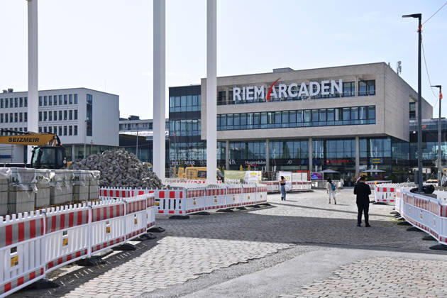 Umgestaltung des Willy-Brandt-Platzes an den Riem Arcaden: Versiegelte Flaechen werden in Gruenflaechen umgewandelt. Am Willy-Brandt-Platz vor den Riem Arcaden in Muenchen laufen die Bauarbeiten zur Neugestaltung. Im Zuge der Umgestaltung sollen bisher versiegelte Flaechen entsiegelt und durch Gruenflaechen ersetzt werden. Baustellenabsperrungen, Baumaterial und ein Radlader praegen derzeit das Bild des Platzes.