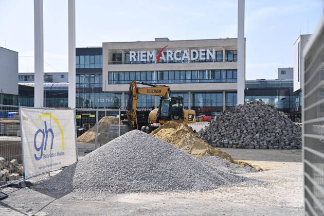 Umgestaltung des Willy-Brandt-Platzes an den Riem Arcaden: Versiegelte Flaechen werden in Gruenflaechen umgewandelt. Am Willy-Brandt-Platz vor den Riem Arcaden in Muenchen laufen die Bauarbeiten zur Neugestaltung. Im Zuge der Umgestaltung sollen bisher versiegelte Flaechen entsiegelt und durch Gruenflaechen ersetzt werden. Baustellenabsperrungen, Baumaterial und ein Radlader praegen derzeit das Bild des Platzes.