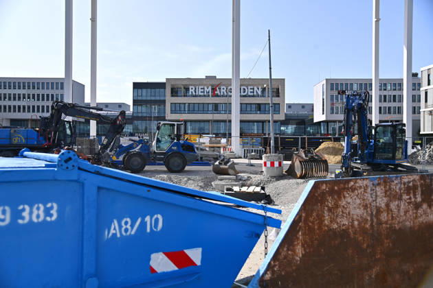 Umgestaltung des Willy-Brandt-Platzes an den Riem Arcaden: Versiegelte Flaechen werden in Gruenflaechen umgewandelt. Am Willy-Brandt-Platz vor den Riem Arcaden in Muenchen laufen die Bauarbeiten zur Neugestaltung. Im Zuge der Umgestaltung sollen bisher versiegelte Flaechen entsiegelt und durch Gruenflaechen ersetzt werden. Baustellenabsperrungen, Baumaterial und ein Radlader praegen derzeit das Bild des Platzes.