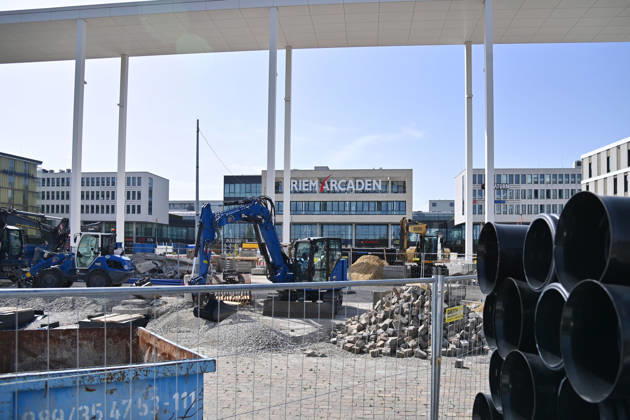 Umgestaltung des Willy-Brandt-Platzes an den Riem Arcaden: Versiegelte Flaechen werden in Gruenflaechen umgewandelt. Am Willy-Brandt-Platz vor den Riem Arcaden in Muenchen laufen die Bauarbeiten zur Neugestaltung. Im Zuge der Umgestaltung sollen bisher versiegelte Flaechen entsiegelt und durch Gruenflaechen ersetzt werden. Baustellenabsperrungen, Baumaterial und ein Radlader praegen derzeit das Bild des Platzes.