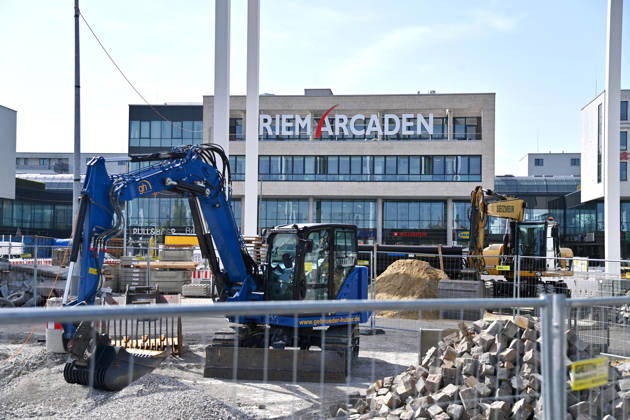 Umgestaltung des Willy-Brandt-Platzes an den Riem Arcaden: Versiegelte Flaechen werden in Gruenflaechen umgewandelt. Am Willy-Brandt-Platz vor den Riem Arcaden in Muenchen laufen die Bauarbeiten zur Neugestaltung. Im Zuge der Umgestaltung sollen bisher versiegelte Flaechen entsiegelt und durch Gruenflaechen ersetzt werden. Baustellenabsperrungen, Baumaterial und ein Radlader praegen derzeit das Bild des Platzes.
