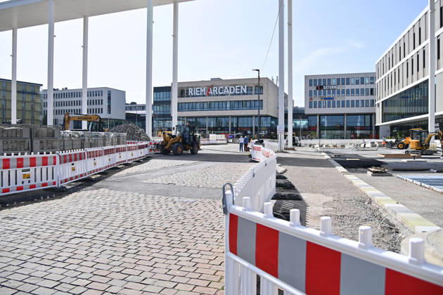 Umgestaltung des Willy-Brandt-Platzes an den Riem Arcaden: Versiegelte Flaechen werden in Gruenflaechen umgewandelt. Am Willy-Brandt-Platz vor den Riem Arcaden in Muenchen laufen die Bauarbeiten zur Neugestaltung. Im Zuge der Umgestaltung sollen bisher versiegelte Flaechen entsiegelt und durch Gruenflaechen ersetzt werden. Baustellenabsperrungen, Baumaterial und ein Radlader praegen derzeit das Bild des Platzes.