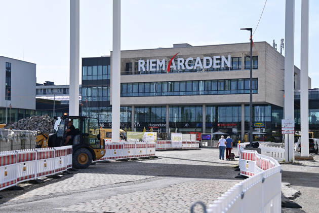 Umgestaltung des Willy-Brandt-Platzes an den Riem Arcaden: Versiegelte Flaechen werden in Gruenflaechen umgewandelt. Am Willy-Brandt-Platz vor den Riem Arcaden in Muenchen laufen die Bauarbeiten zur Neugestaltung. Im Zuge der Umgestaltung sollen bisher versiegelte Flaechen entsiegelt und durch Gruenflaechen ersetzt werden. Baustellenabsperrungen, Baumaterial und ein Radlader praegen derzeit das Bild des Platzes.