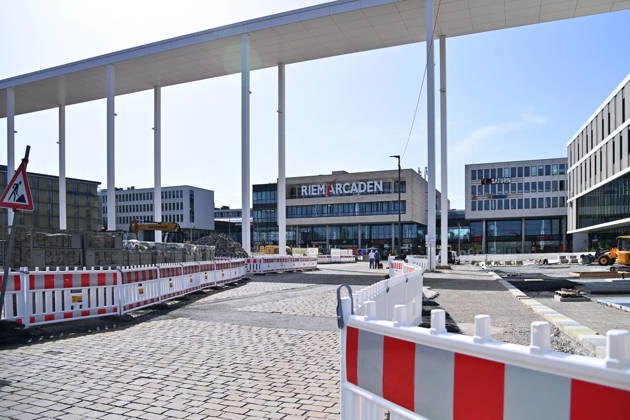 Umgestaltung des Willy-Brandt-Platzes an den Riem Arcaden: Versiegelte Flaechen werden in Gruenflaechen umgewandelt. Am Willy-Brandt-Platz vor den Riem Arcaden in Muenchen laufen die Bauarbeiten zur Neugestaltung. Im Zuge der Umgestaltung sollen bisher versiegelte Flaechen entsiegelt und durch Gruenflaechen ersetzt werden. Baustellenabsperrungen, Baumaterial und ein Radlader praegen derzeit das Bild des Platzes.