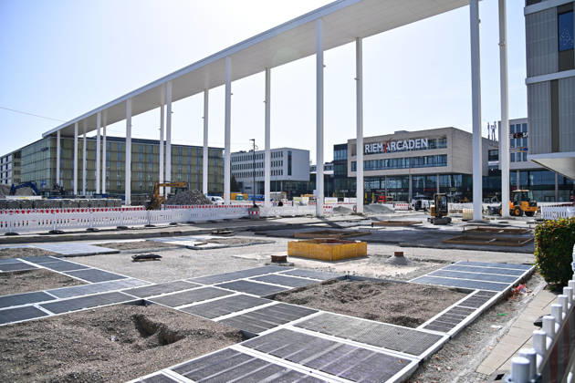 Umgestaltung des Willy-Brandt-Platzes an den Riem Arcaden: Versiegelte Flaechen werden in Gruenflaechen umgewandelt. Am Willy-Brandt-Platz vor den Riem Arcaden in Muenchen laufen die Bauarbeiten zur Neugestaltung. Im Zuge der Umgestaltung sollen bisher versiegelte Flaechen entsiegelt und durch Gruenflaechen ersetzt werden. Baustellenabsperrungen, Baumaterial und ein Radlader praegen derzeit das Bild des Platzes.