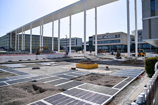 Umgestaltung des Willy-Brandt-Platzes an den Riem Arcaden: Versiegelte Flaechen werden in Gruenflaechen umgewandelt. Am Willy-Brandt-Platz vor den Riem Arcaden in Muenchen laufen die Bauarbeiten zur Neugestaltung. Im Zuge der Umgestaltung sollen bisher versiegelte Flaechen entsiegelt und durch Gruenflaechen ersetzt werden. Baustellenabsperrungen, Baumaterial und ein Radlader praegen derzeit das Bild des Platzes.