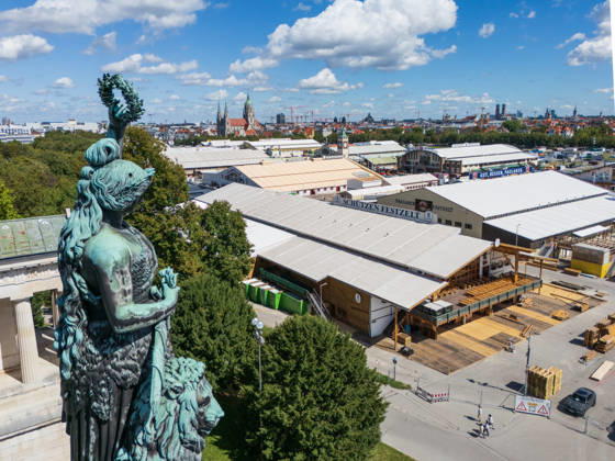 MUNICH, GERMANY - AUGUST 29, 2026: Bavaria Statue Overlooking Oktoberfest Setup The Bavaria statue stands beside its lion, overlooking the Theresienwiese during the setup phase of the 2026 Oktoberfest. Below, large beer tents such as the Schuetzen Festzelt and Paulaner rise among wooden structures and open festival grounds, with Munich s skyline visible under a bright summer sky.