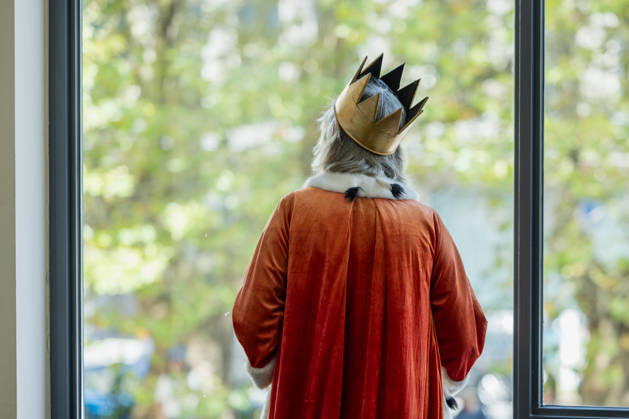 Kostümparade derr Deutschen Opar Berlin. Deutschland, Berlin am 29. August 2026: Mit dem Slogan Unless the People Live Here eröffnete die Deutsche Oper Berlin die neue Spielzeit bzw Saison. Zahlreiche Besucherinnen und Besucher verkleideten sich mit Kostümen aus dem Fundus der Oper. Eine als König verkleidete Person mit grauen Haaren, goldener Krone und rotem Hermelinumhang blickt aus einem Fenster.