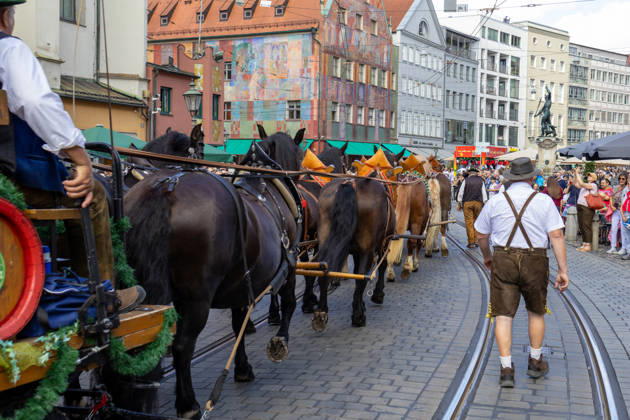 Traditioneller Plärrerumzug in Augsburg. Ein traditionelles Pferdegespann mit geschmückten Kaltblütern und Festwagen zieht beim Plärrerumzug durch die Augsburger Innenstadt. Zahlreiche Zuschauer säumen die Strecke des Festzugs