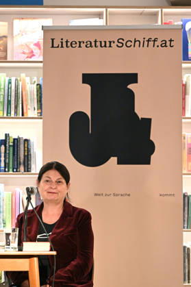 Minifestival AH0J (Literaturfestival) - Tschechien setzt über - der Kulturinitiative Literaturschiff, in der Buchhandlung Kurdirektion in Bad Ischl. Lesungen und Gespräche in Korrespondenz zum Gastauftritt Tschechiens auf der Frankfurter Buchmesse 2026. Es liest die tschechische Schriftstellerin und mehrfache Literaturpreisträgerin Radka Denemarkovich. Die Veranstaltung wrd moderiert von Julia Miesenböck, fotografiert am 30.09.26. Das Bild zei vor einem Plakat der Kulturinitiative Literaturschiffgt die Schriftstellerin Radka Denemarkovic 2026 - Minifestival AH0J (Literaturfestival) - Tschechien setzt über - der Kulturinitiative Literaturschiff, in der Buchhandlung Kurdirektion in Bad Ischl. Lesungen und Gespräche in Korrespondenz zum