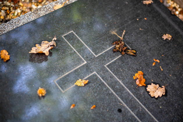 Auf einer dunklen, polierten Grabplatte mit einem eingravierten Kreuz liegen vereinzelte, braune Eichenblätter im herbstlichen Licht. Rund um Eicks